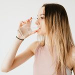Young thirsty fit female with long hair in sportswear drinking water while recreating after workout